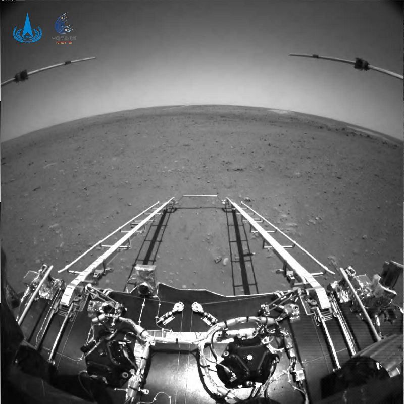 △由火星车前避障相机拍摄，正对火星车前进方向。图中可见坡道机构展开正常；图像上部的两个伸杆为已经展开到位的次表层雷达；前进方向地形清晰。为获知火星车前进方向更大范围的地形信息，避障相机采用大广角镜头，在广角镜头畸变的影响下，远处地平线形成一条弧线。