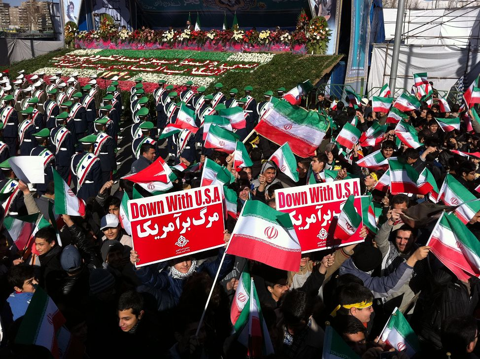 伊朗2012年革命日聚会现场，少年舞动着“打倒美国”的标语，远处主席台是时任总统内贾德的演讲。王文 摄