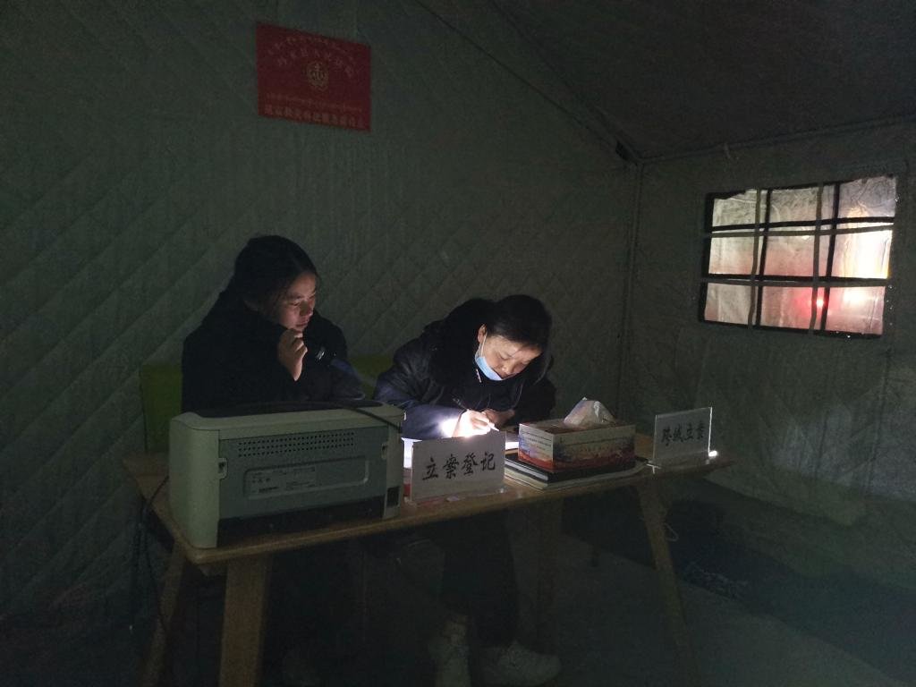 2021年5月26日，在抗震救灾临时办公点的帐篷内，玛多县人民法院法官助理更周措（右）在手电筒照射的灯光下进行立案登记。受访者供图