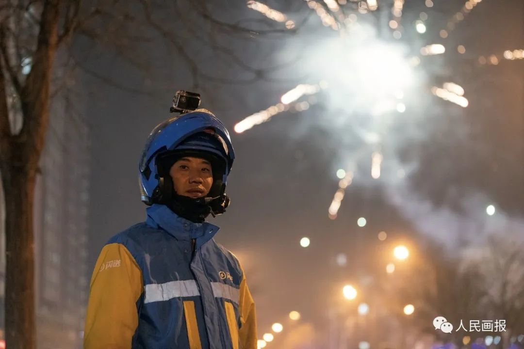 2021年2月11日,农历大年三十,北京市朝阳区,闪送员许凌志送完一单业务后在街边休息,等待下一单业务。此时已临近午夜12点,远处璀璨的焰火预示着新年的到来。许凌志响应国家就地过年的号召,留在北京过春节。“忙,能赚钱,还能帮到别人,挺好。”许凌志说。摄影 段崴/人民画报