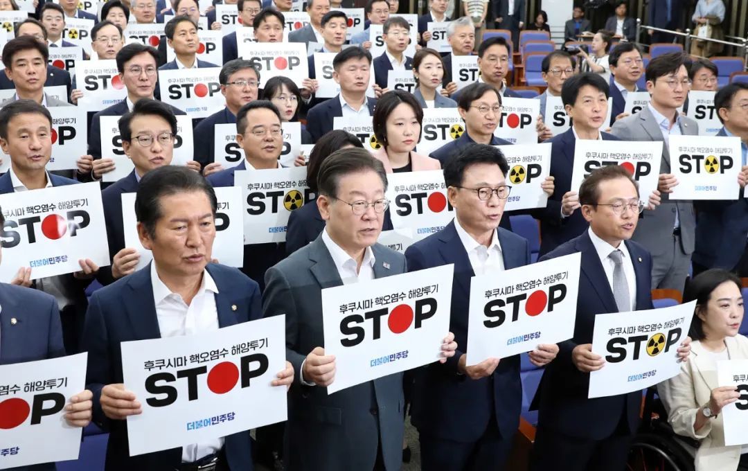 8月24日，韩国最大在野党共同民主党党首李在明（前排左二）与议员在首尔国会举行的紧急会议上手举标语，抗议日本核污染水排海 / 图源：新华社