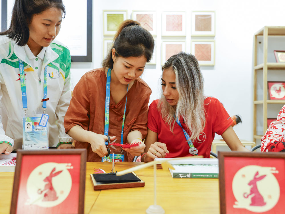 8月2日,成都大运村互动体验中心,非遗项目青神竹编代表性传承人赵爽(中)在指导土耳其运动员塞姆拉(右)制作竹编。新华社记者沈伯韩摄