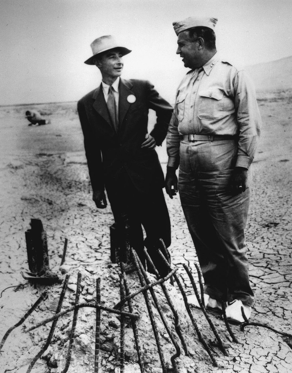 1945年9月，奥本海默（左）和莱斯利·格罗夫斯将军在“三位一体”核弹试验场遗址。此时三位一体核试验已过去两个月，二战刚刚结束不久。他们穿着白色的鞋套，防止放射性尘埃沾到鞋底。© Wikimedia Commons