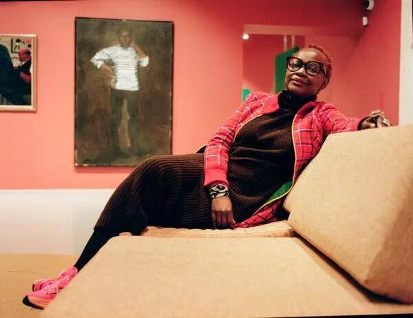 A woman wearing a colorful jacket sits on a couch in front of a painting of a Black man