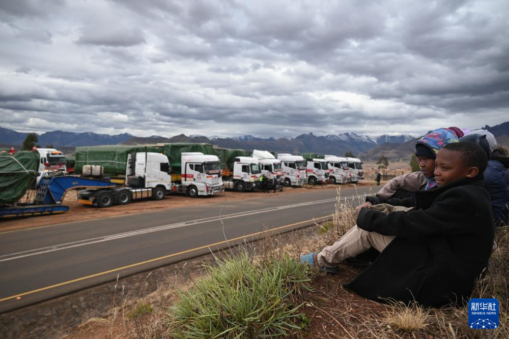 7月6日，在位于莱索托北部的莱里贝地区皮岑附近的山区公路旁，当地儿童席地而坐围观运送硬岩掘进机部件的卡车车队。新华社记者 杨光 摄
