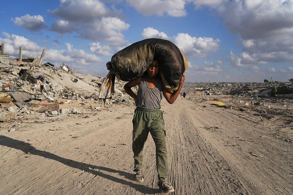 当地时间2025年10月12日,加沙城,以色列和哈马斯同意暂停冲突并释放剩余人质后,一名流离失所的巴勒斯坦男孩带着自己的物品走在满目疮痍的街道。视觉中国 图