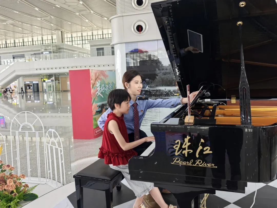 在人来人往的车站内,一位小旅客正与工作人员在音乐驿站合奏钢琴曲。