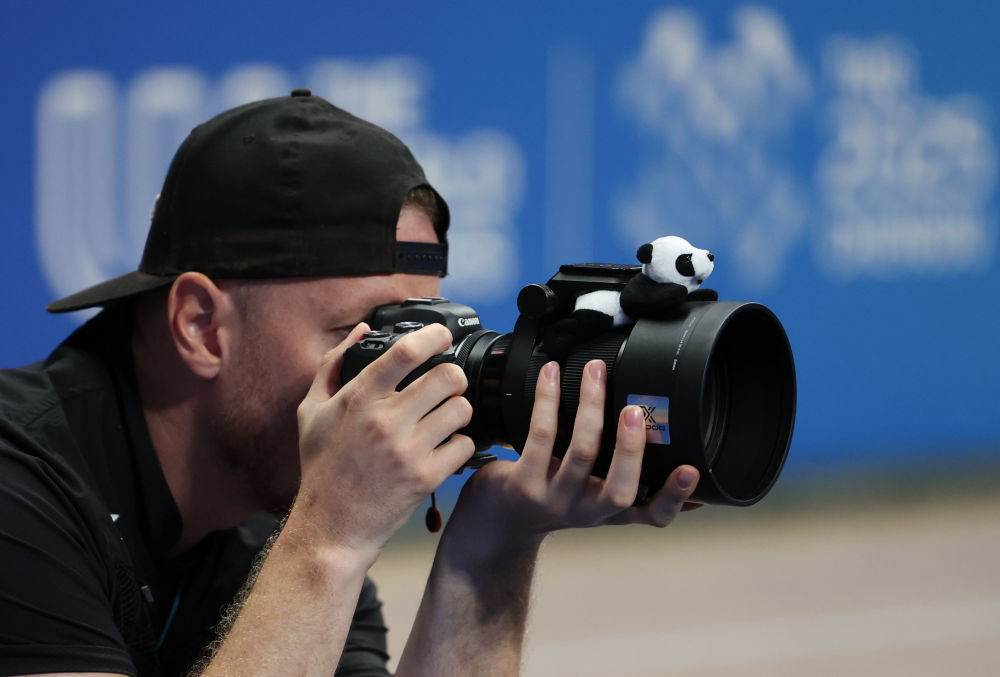 ↑8月13日,一名外媒记者在成都世运会软式曲棍球男子铜牌赛拍摄,他的相机镜头上装饰了熊猫玩偶。