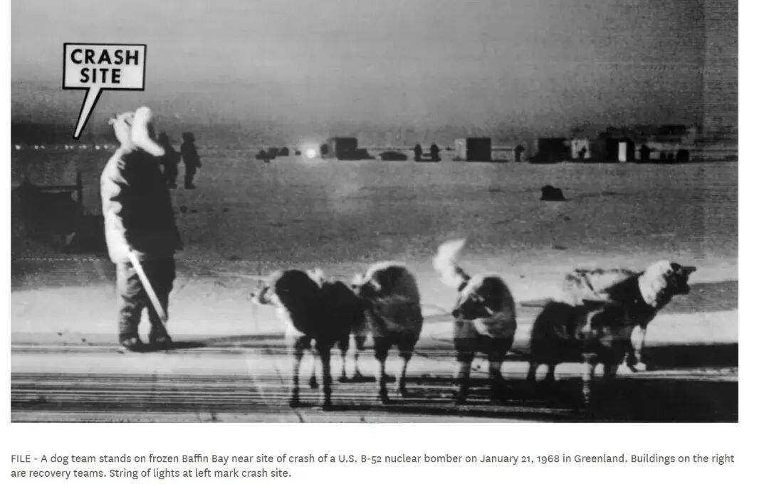 1968年1月21日，在格陵兰，一队雪橇犬站在结冰的巴芬湾，靠近一架美国B-52核轰炸机的坠毁地点。右侧的建筑是回收（打捞）队伍的驻地，左侧的一串灯光标记着坠机位置。