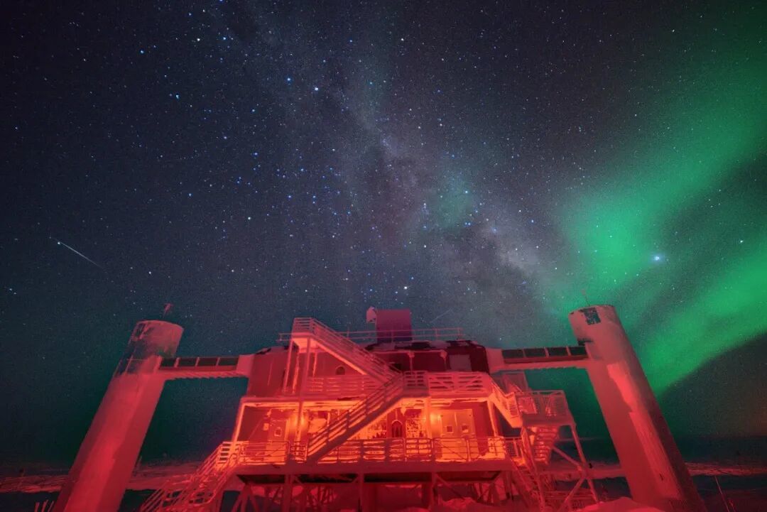 南极冬季夜空下的冰立方实验室,头顶是银河与极光,探测器就埋在它下方的冰层里。丨John Hardin/Wikimedia Commons(CC BY 4.0)