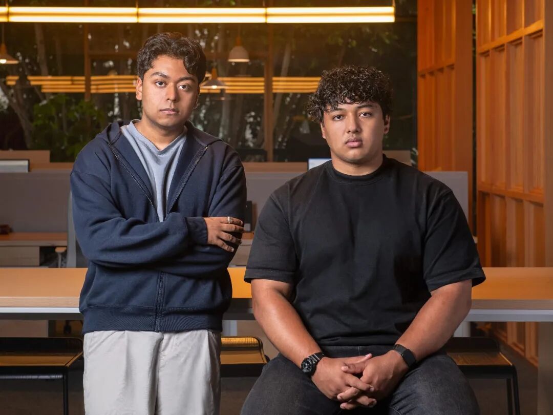 Architect Labs cofounders Ebrahim Hussain and Aaditya Subedi, in front of a desk with flourescent lights and wood walls.