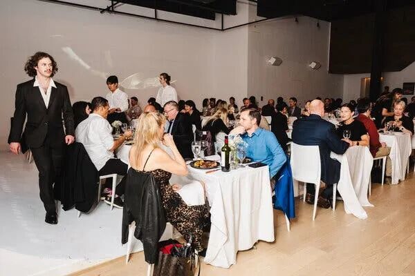 Diners sit for dinner in an event space turned into a dining room, complete with white tablecloths and servers in suits.