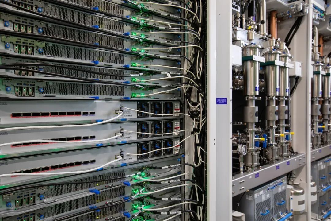 Close-up of server racks with connected cables and green indicator lights beside metal piping and cooling equipment.