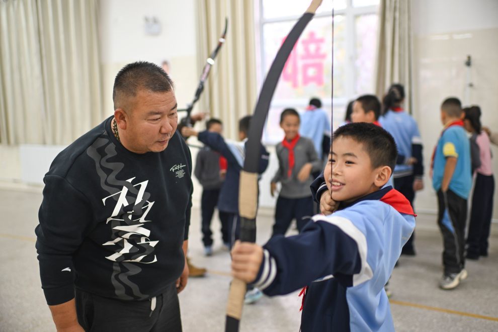 在新疆伊宁市第二十五小学,非遗传承人吴俊亮(左)指导射箭社团学生练习传统弓(2025年10月23日摄)。新华社记者 胡虎虎 摄