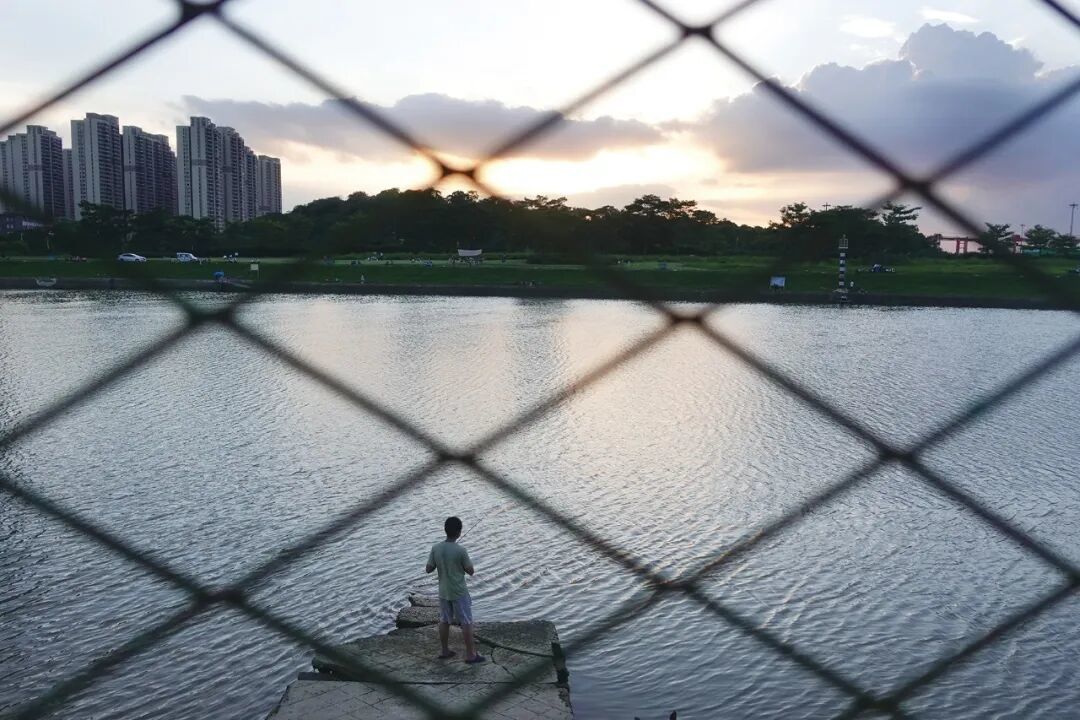 2023年9月17日,广东广州。黄昏时分,一名男子独自在江边钓鱼。照片中的人恰好落在菱形网格之中,形成对当代生活的一种隐喻。(图/苏峥)
