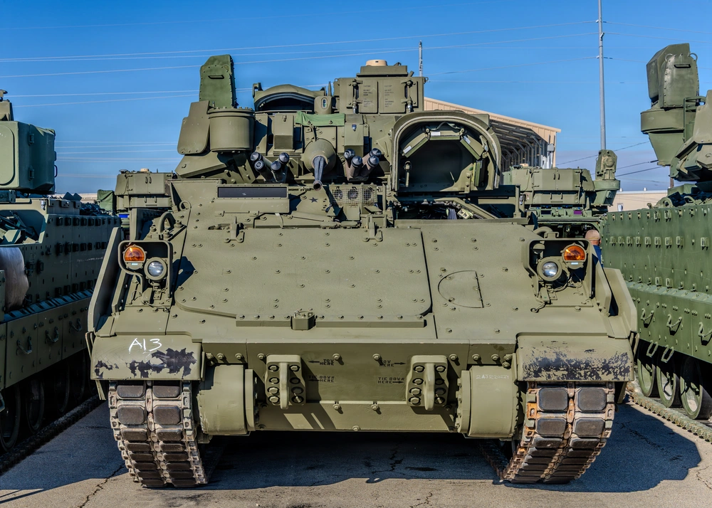A front-facing view of a U.S. Army Bradley Fighting Vehicle (BFV) marked 'A13' in white chalk on its lower left hull. The vehicle is painted in olive drab and is parked among other similar armored vehicles in what appears to be a military staging or storage yard. The image is taken in daylight with clear skies, showing considerable detail of the vehicle's front armor, tracks, and turret assembly.
