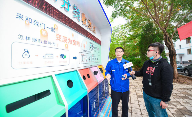 金昌市环保小屋回收管家（左）介绍再生资源收集点智能垃圾分类箱使用情况