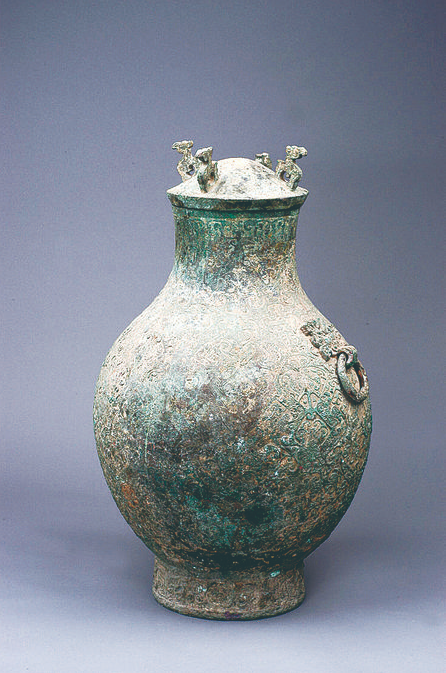 ▲大铜壶 礼器。口径16厘米、腹35厘米、高57厘米。拱状,圆顶4环状,龙鸟纹盖系,下有牙子盖,壶体平口唇,长颈,圆鼓腹,高商圈足,肩部有两铺首饰环,其一已失。空间缀饰以花叶纹,勾连纹饰近底部有一圈宽固带纹,高圈足部有两道带纹,其中施以窃曲,成组云卷纹。