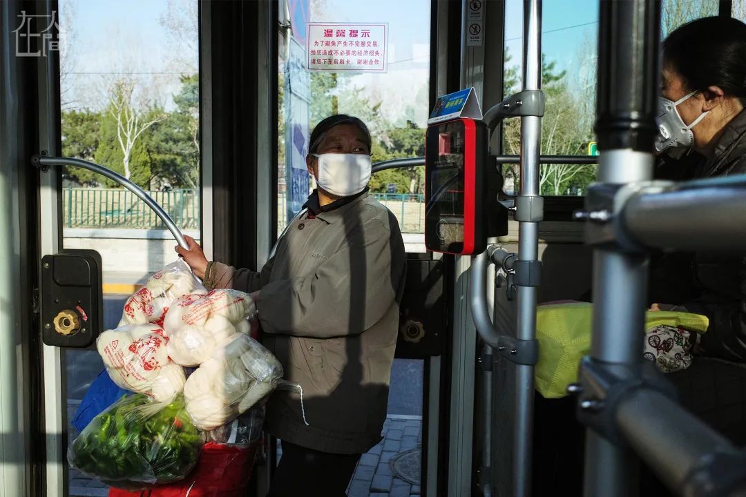 ▲ 3月17日，北京。公交车上，一位老人正在向一旁的乘客展示自己因疫情准备的蔬菜和馒头。