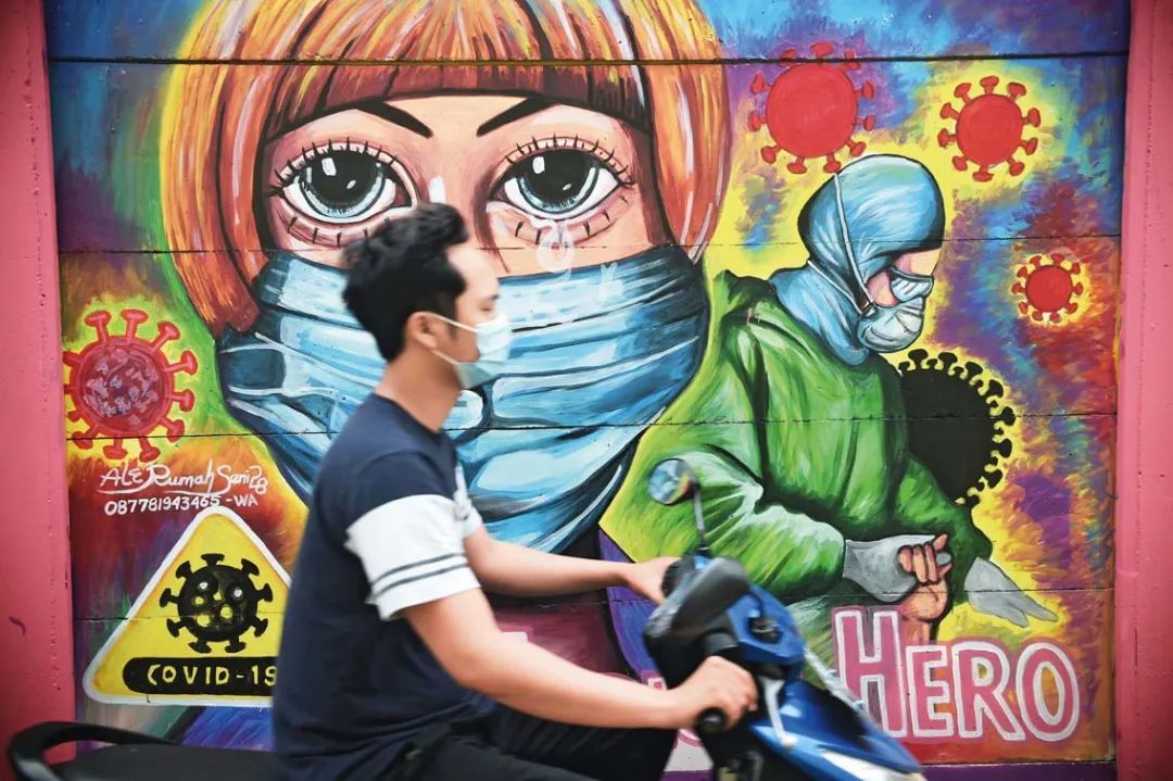2021 年 1 月 26 日，在印度尼西亚唐格朗，一名男子骑摩托车从一处描绘新冠肺炎疫情的壁画前驶过
