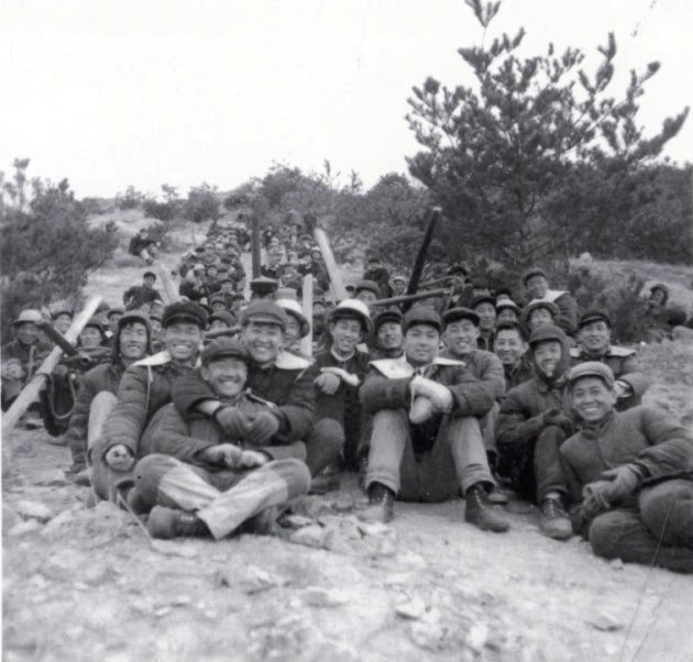 (1964年3月,第六地质大队探矿人员在九曲矿区钻探施工,当时运送钻探设备全靠肩扛人抬。此图为寒风刺骨,滴水成冰的冬季,组织机关及一线人员100余人往20多度陡坡上搬运设备休息时的场景。)