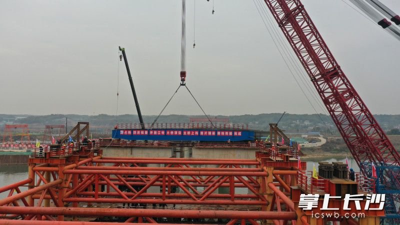 目前,暮坪湘江特大桥项目正处在主体冲刺施工阶段。该项目是S326浏阳澄潭江至宁乡沙田公路工程(长沙市南横线)的过江通道,连接天心区南部片区与湘江新区大王山片区。项目河东起点位于万家丽路与中意路交叉口以西约800米处,向西依次跨越京广铁路、湘江大道、湘江和潇湘大道,河西终点位干坪塘大道以东约768米处,线路全长约4.416公里。主桥为四跨中承飞燕式钢桁系杆拱桥,宽42.5米;东、西涉水引桥为钢-混组合梁桥,桥宽38.5米。
