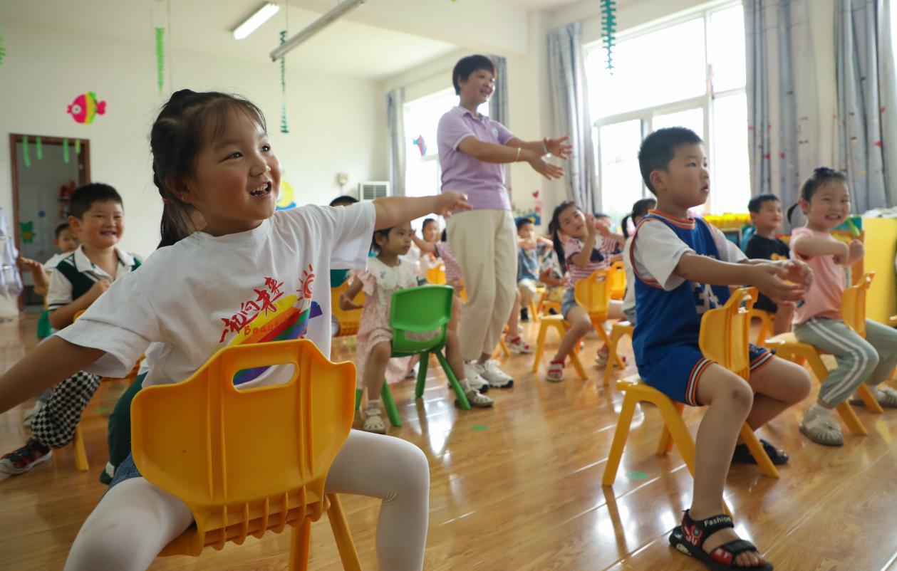 （2023年7月13日，在安徽省合肥市肥西县山南镇中心幼儿园，孩子们在老师的指导下学习椅子舞。 陈家乐 摄）
