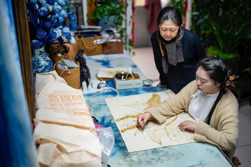 ↑4月6日,李萨(右)在工作室内为顾客演示蜡染工艺品上蜡。
