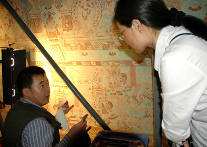 为了让游客了解世界文化遗产敦煌莫高窟的壁画保存现状、保护原则、保护方式，提高公众对莫高窟文物的保护意识，2008年6月以来，敦煌莫高窟壁画保护修复现场每周定期向游客开放。图为2008年9月22日，在莫高窟98窟内，敦煌研究院保护人员（左）正在向一名游客讲解如何修复壁画。新华社记者　王艳明　摄