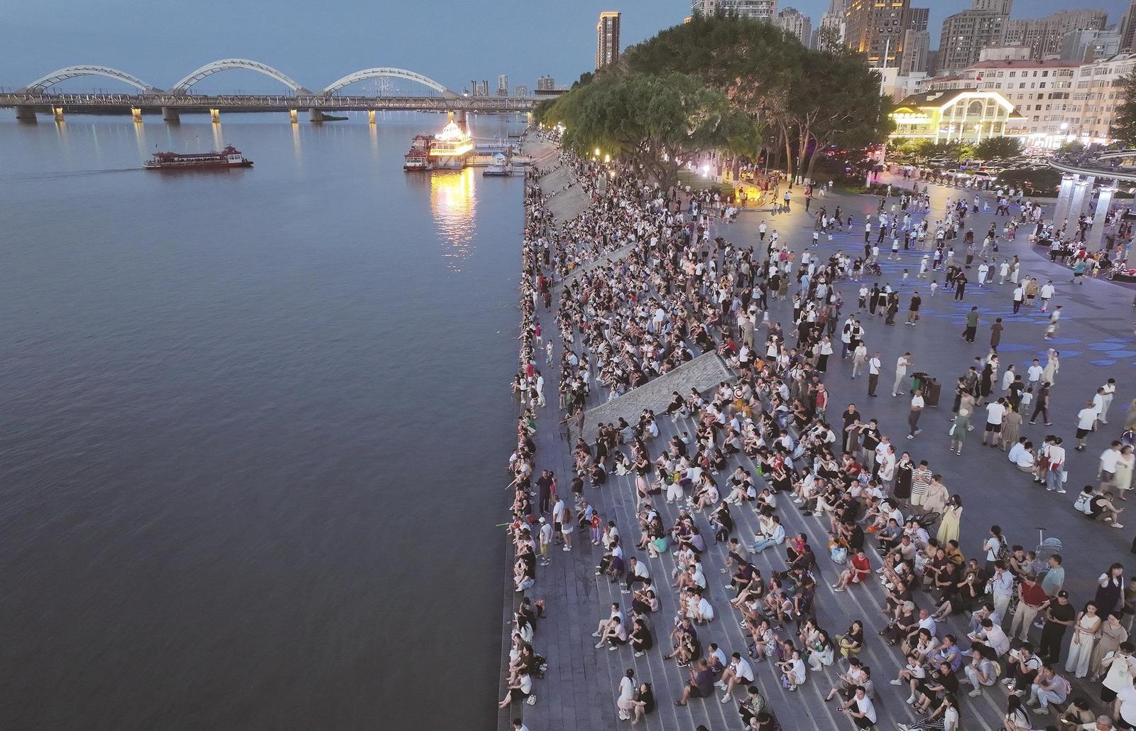 7月25日，在哈尔滨市防洪纪念塔前的江边，人们坐在台阶上休闲纳凉，观赏江景（无人机照片）。新华社发 张树 摄