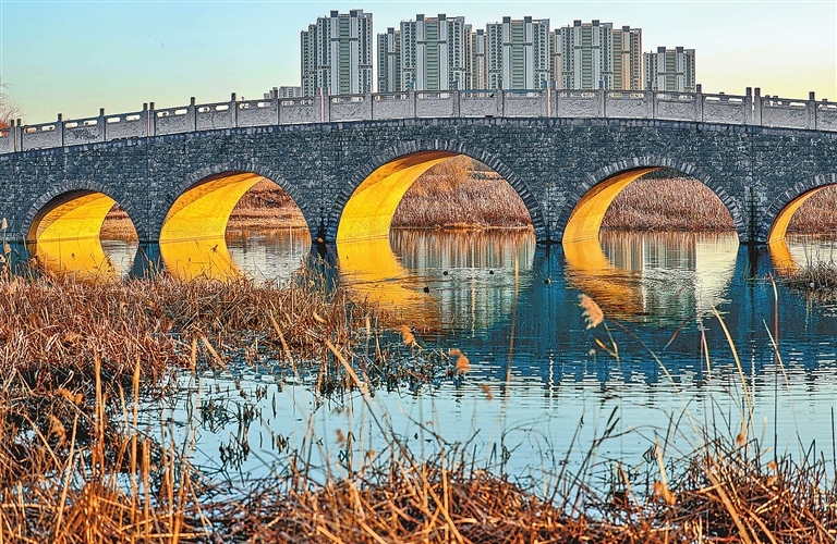 2025年12月14日，在郑州市陇海西路与丹河大道旁的贾鲁河上，一座拱桥的桥洞被金光填满，其倒影在波光粼粼的水面舒展铺陈。
