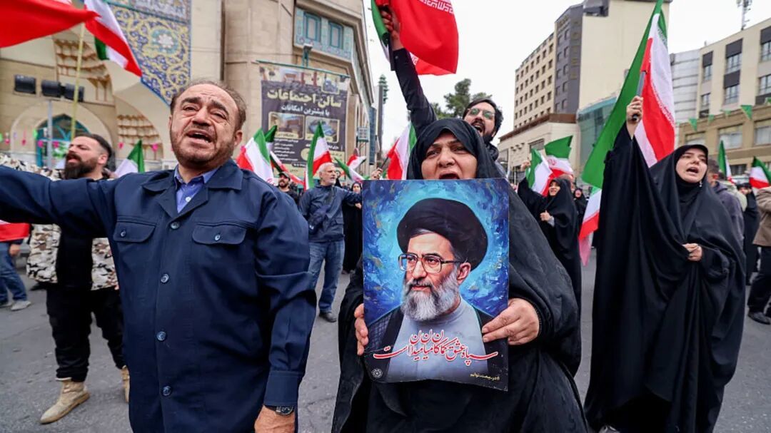 一名女性手持一幅描绘伊朗最高阿亚图拉&middot;阿里&middot;哈梅内伊的插画，2026年2月28日，其他人在德黑兰一座清真寺外举行支持政府、反对美以空袭的示威中挥舞伊朗国旗。