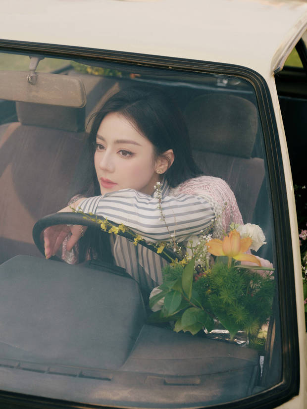 迪丽热巴手搭方向盘，身旁鲜花环绕，眼神温柔，治愈浪漫。